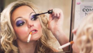 A blonde woman applying eye makeup in a mirror while smoking