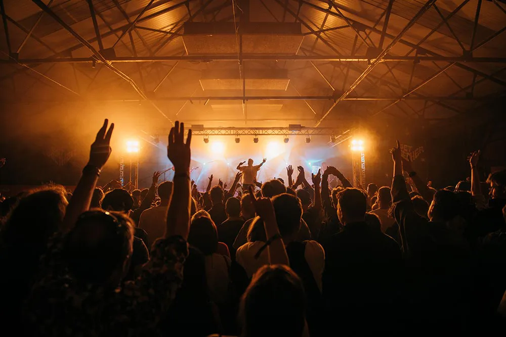 A crowd stand in a darkly lit room in front of a stage with one performer.